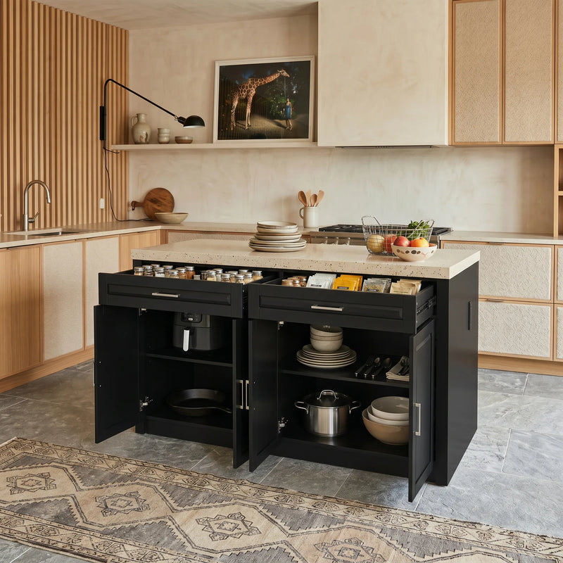 A modern kitchen island cabinet in matte black, shown open to reveal storage compartments and drawers. The kitchen features warm beige wood and textured cabinets, a light stone countertop, and gray stone tile flooring with a patterned rug.