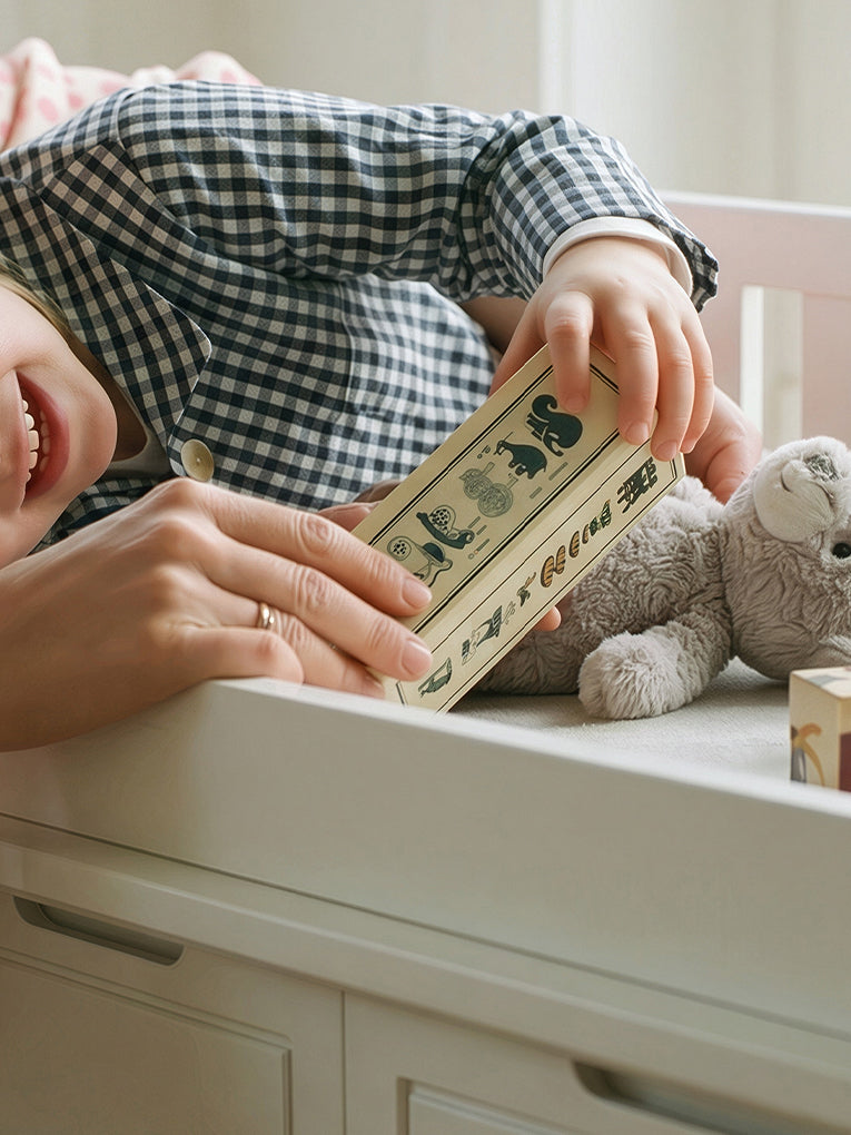 A close-up lifestyle shot of a child in a blue and white gingham pajama shirt holding a wooden toy block with animal illustrations, with an adult's hand guiding them, next to a gray plush toy on a white dresser.