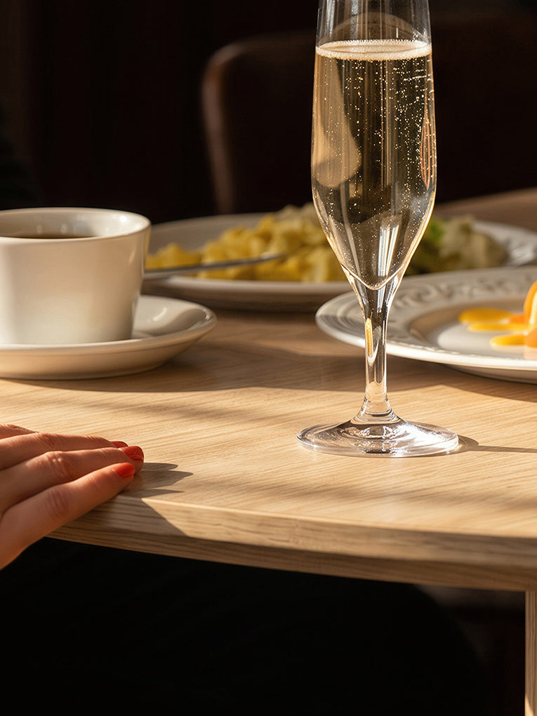 A clear champagne flute filled with bubbly golden liquid sits on a light wood table in warm sunlight, with a white cup and plates blurred in the background. A hand with red nail polish rests at the left edge.
