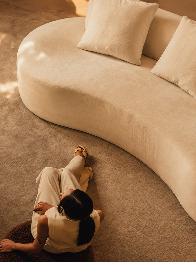 A woman sits on the floor in front of a large, curved cream-colored sofa with matching throw pillows, viewed from an overhead angle in a warm, minimalist interior.