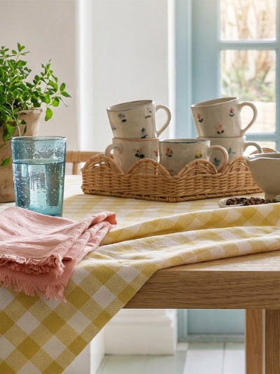 A yellow and white gingham check tablecloth draped over a wooden dining table, styled with a pink fringed napkin, a blue glass, and a wicker tray holding cream-colored floral ceramic mugs.