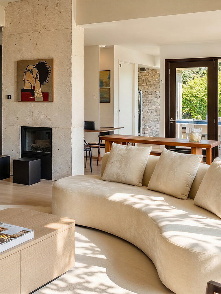 A modern curved beige sofa in a bright, contemporary living room with a stone fireplace, wooden dining table, and large windows.