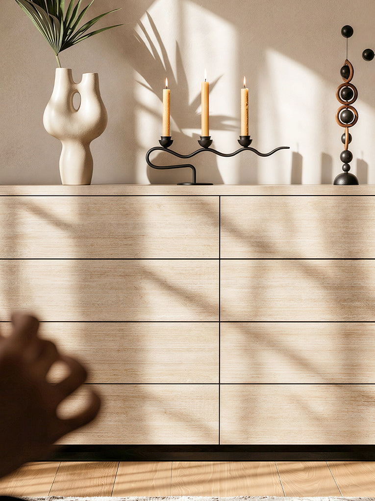 A modern light wood console cabinet in a warm beige tone, styled with a white vase, candles, and decorative objects on top, set against a beige wall with soft window shadow patterns.