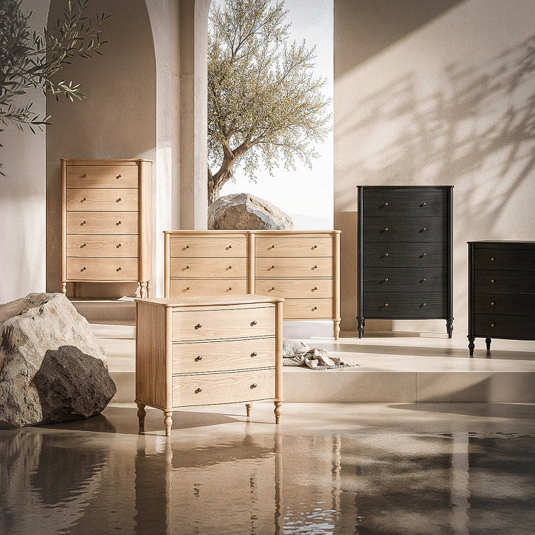 A collection of wooden chest of drawers in light natural oak and dark espresso finishes, arranged in a sunlit interior setting with stone columns, rocks, and a tree visible in the background.