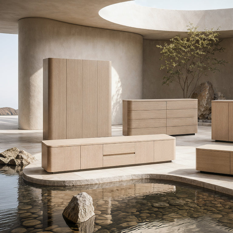 A minimalist furniture set in light natural wood tones arranged in a serene architectural interior with curved concrete walls, a tree, rocks, and a reflective water feature in the foreground.