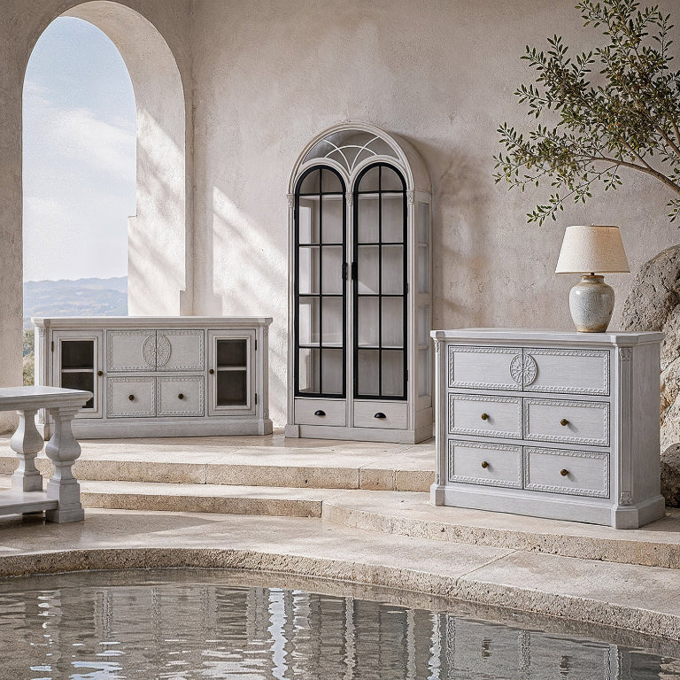 A coordinated set of light gray carved wooden furniture pieces arranged in a sunlit stone interior beside a pool, including a tall arched glass-front cabinet, a sideboard, a chest of drawers, and a table lamp.